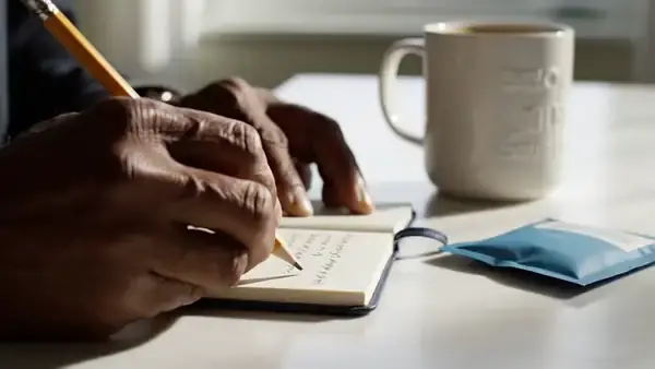 A man's hands writing a line and a check mark in a small notebook beside a mug of coffee and a blue pouch